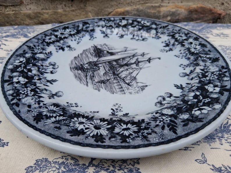 Pu&ograve; includere: Piatto in ceramica bianca con bordo floreale blu e bianco e un'illustrazione centrale di una nave. Il piatto ha un'estetica classica e vintage, adatto per servire cibo o come elemento decorativo.