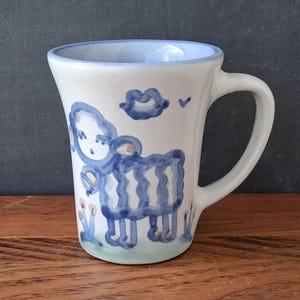 May include: A white ceramic mug featuring a hand-painted blue sheep illustration on the front. The design includes a small blue cloud shape above the sheep and simple blue floral accents at the base. The mug has a flared rim and a curved handle.