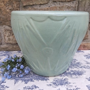 Könnte beinhalten: Ein großer, hellgrüner Keramik-Pflanztopf mit einem strukturierten, blattartigen Design. Der Topf steht auf einem blau-weiß gemusterten Stoff, mit einem kleinen Strauß blauer Blumen links. Der Hintergrund ist eine Steinmauer.