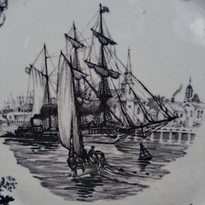 Pu&ograve; includere: Piatto bianco e blu con un'illustrazione in bianco e nero di una nave a vela in un porto. La scena comprende altre navi, edifici e un bordo floreale. Il piatto ha un design vintage.