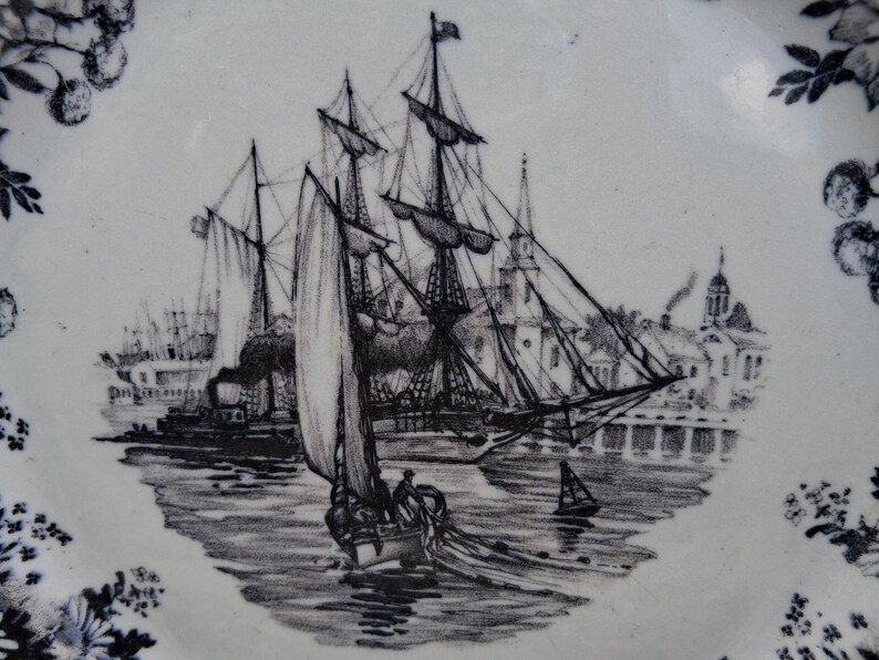 Pu&ograve; includere: Piatto bianco e blu con un'illustrazione in bianco e nero di una nave a vela in un porto. La scena comprende altre navi, edifici e un bordo floreale. Il piatto ha un design vintage.