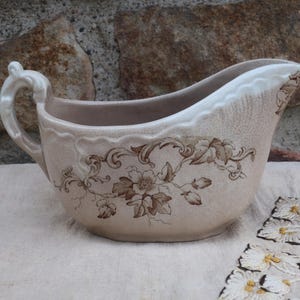 以下が含まれることがあります： 茶色の花柄が施された白い陶器製のグレービーボート。ボートにはハンドルと注ぎ口があります。