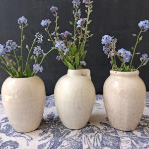 May include: Three off-white ceramic vases, each holding sprigs of blue forget-me-not flowers. The vases have a crackled glaze finish and varying shapes. The arrangement sits on a blue and white patterned fabric against a dark background.