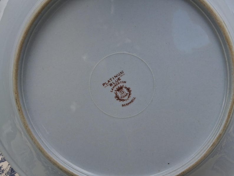 Pu&ograve; includere: Un piatto in ceramica azzurra con bordo marrone. Il centro del piatto presenta un emblema circolare con la scritta "PLATINUM BLUE LAMBERTON CHINA SCAMMELL".