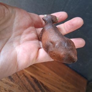 May include: A small, brown, textured figurine of a four-legged animal, possibly a cow or bull, featuring a smooth, rounded body, a distinct head with small horns, and a dark, earthy finish that highlights the sculpted contours of the creature.