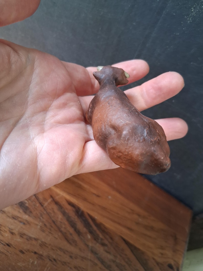 May include: A small, brown, textured figurine of a four-legged animal, possibly a cow or bull, featuring a smooth, rounded body, a distinct head with small horns, and a dark, earthy finish that highlights the sculpted contours of the creature.