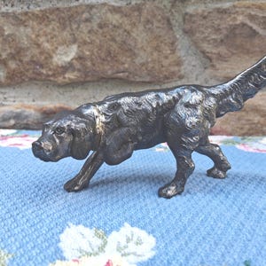 May include: A bronze-colored dog figurine in a walking pose. The sculpture has a textured surface and is set against a blue floral patterned fabric and a stone background. The dog's tail is raised.