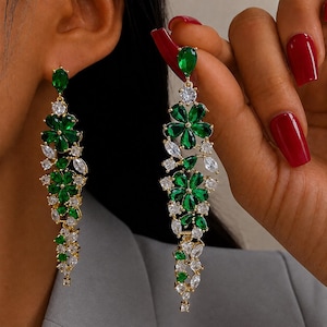 Peut inclure: Longues boucles d'oreilles pendantes dorées au design floral en cascade. Chaque boucle est ornée de multiples cristaux vert émeraude en forme de poire et de cristaux blancs transparents ronds et en marquise sur une monture dorée.