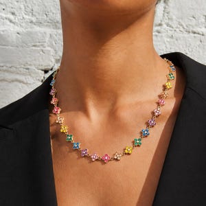Könnte beinhalten: Eine goldfarbene Halskette mit einer Reihe kleiner, vierblättriger Blumenanhänger. Jeder Blumenanhänger ist mit winzigen, leuchtend bunten Edelsteinen in den Farben Blau, Grün, Gelb, Pink und Violett besetzt, die einen Regenbogeneffekt erzeugen.