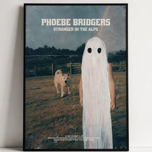 Puede incluir: Un póster enmarcado con el texto PHOEBE BRIDGERS STRANGER IN THE ALPS. La imagen muestra una figura cubierta con una sábana blanca con dos agujeros negros para los ojos, de pie en un campo junto a un perro claro, con un cielo oscuro y una valla de madera.