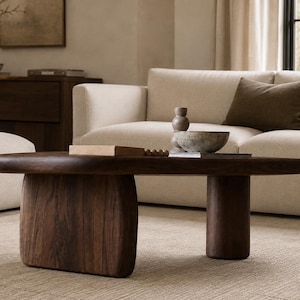 May include: A low-profile coffee table crafted from dark brown wood with a smooth, rounded oval top. The table is supported by two distinct legs: one wide, flat vertical panel and one thick, cylindrical pillar, both featuring a visible wood grain.