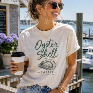 May include: A cream-coloured short-sleeve t-shirt featuring a green graphic print that reads Oyster Shell, with an illustration of an oyster shell and the text COFFEE WELLFLEET, MA below it. The shirt has a crew neckline and rolled sleeves.