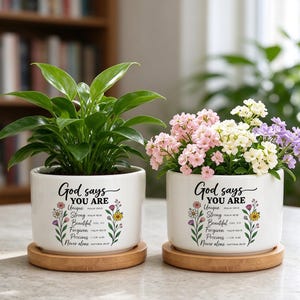 May include: Two white ceramic cylindrical planters on round wooden saucers. Each planter features the black text God says YOU ARE with a list of attributes and floral illustrations. One contains a green plant, the other pink and white flowers.