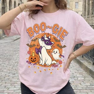 Puede incluir: Una camiseta rosa claro con un gráfico de un gato blanco con máscara y pajarita moradas, un pequeño fantasma, una calabaza de Halloween y caramelos. El texto "BOO-GIE" está impreso en naranja con borde blanco sobre un círculo naranja.