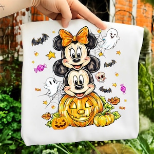 May include: A white t-shirt featuring a colorful Halloween graphic of Mickey and Minnie Mouse stacked on top of a large jack-o'-lantern. The design includes small ghosts, bats, candy corn, wrapped candies, a skull, and spider webs.
