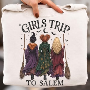 May include: A cream-colored sweatshirt featuring a graphic of three figures from behind, each holding a broomstick. The text GIRLS TRIP TO SALEM is printed above and below the figures, accompanied by illustrations of stars, bats, and a crescent moon.