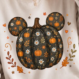 Könnte beinhalten: Ein weißes Sweatshirt mit Rundhalsausschnitt und einem großen Aufdruck eines dunkelgrauen Kürbisses in Form von Mausohren. Der Kürbis ist mit kleinen orangefarbenen Kürbissen, weißen Gänseblümchen und braunen Blättern verziert. Kleine orangefarbene Herzen, Sterne und Schnörkel umgeben das Motiv.