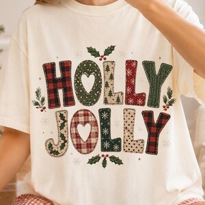 Può includere: Una t-shirt a maniche corte color crema con la scritta HOLLY JOLLY in grandi lettere a motivi vari. Le lettere presentano motivi a quadri rossi e neri, verde con alberi bianchi e beige con fiocchi di neve, decorati con foglie di agrifoglio.