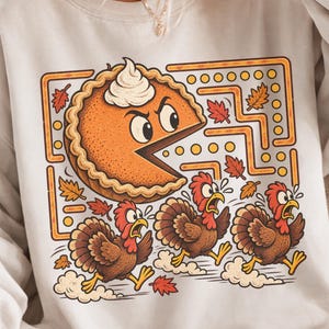 May include: A white crewneck sweatshirt featuring a graphic print of a pumpkin pie slice with a face chasing three brown turkeys through a yellow maze. The design includes scattered orange and red autumn leaves and a dollop of whipped cream.