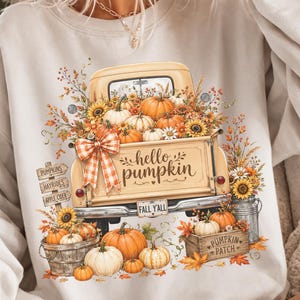 Puede incluir: Una sudadera de cuello redondo de color claro con un gráfico de un camión vintage lleno de calabazas, girasoles y follaje otoñal. El portón trasero del camión muestra el texto "hello pumpkin" y "FALL Y'ALL" junto a un lazo a cuadros.