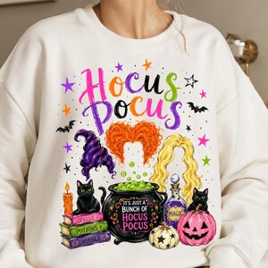 May include: A white crewneck sweatshirt featuring a colourful graphic print with the text Hocus Pocus. The design includes three stylised hairstyles, two black cats, a cauldron, a pumpkin, a candle, and books labelled Spells, Potions, and Trouble.