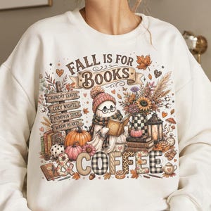 May include: A white long-sleeve sweatshirt featuring a graphic print with the text "FALL IS FOR BOOKS & COFFEE". The design includes a ghost wearing glasses and a beanie reading a book, surrounded by pumpkins, autumn leaves, and a lantern.