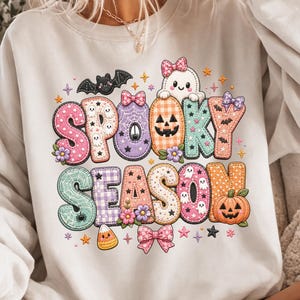 Op de afbeelding: Een wit sweatshirt met lange mouwen en een kleurrijke grafische print met de tekst "SPOOKY SEASON" in een doorgestikt lettertype. Het ontwerp bevat illustraties van een spook, een vleermuis, pompoenen, snoepmaïs en strikjes in pasteltinten.