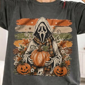 May include: A charcoal gray t-shirt featuring a graphic print of a hooded figure with a ghost-like mask holding a pumpkin. The design includes autumn leaves, flowers, and two jack-o'-lanterns against a background of horizontal stripes.