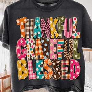 May include: A charcoal gray short-sleeve t-shirt featuring the words THANKFUL GRATEFUL BLESSED in large, block-style letters. Each letter displays a unique pattern, including polka dots, leopard print, plaid, stripes, stars, and floral designs.