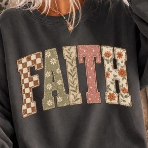 May include: A charcoal gray crewneck sweatshirt featuring the word FAITH printed across the chest. Each letter has a unique pattern: a brown and white checkerboard, sage green with white daisies, green vines, dusty pink with stars, and orange flowers.