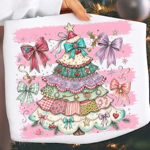 Puede incluir: Una sudadera blanca con un gráfico de un árbol de Navidad de varias capas decorado con varios patrones, incluidos cuadros y lunares. El árbol está adornado con lazos, cerezas y estrellas, rodeado de pinceladas horizontales de color rosa.