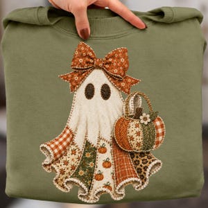 Puede incluir: Una sudadera verde oliva con el gráfico de un fantasma que lleva un lazo grande y sostiene una cesta de calabaza. El fantasma y la calabaza están decorados con varios patrones de patchwork, incluyendo cuadros vichy, flores y estampado de leopardo.