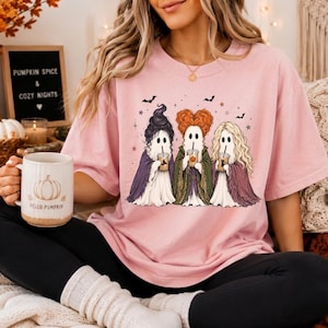 May include: A light pink t-shirt featuring a graphic of three ghosts dressed as witches, each holding a coffee cup. The ghosts have distinct hairstyles in black, orange, and blonde, and are wearing purple, green, and maroon capes with small bats.