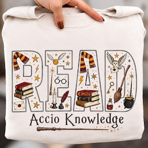 May include: A cream-colored sweatshirt featuring the word READ in large block letters filled with illustrations of books, a scarf, a wand, glasses, a candle, an owl, a broom, and a cauldron. Below, the text Accio Knowledge appears with a wand.