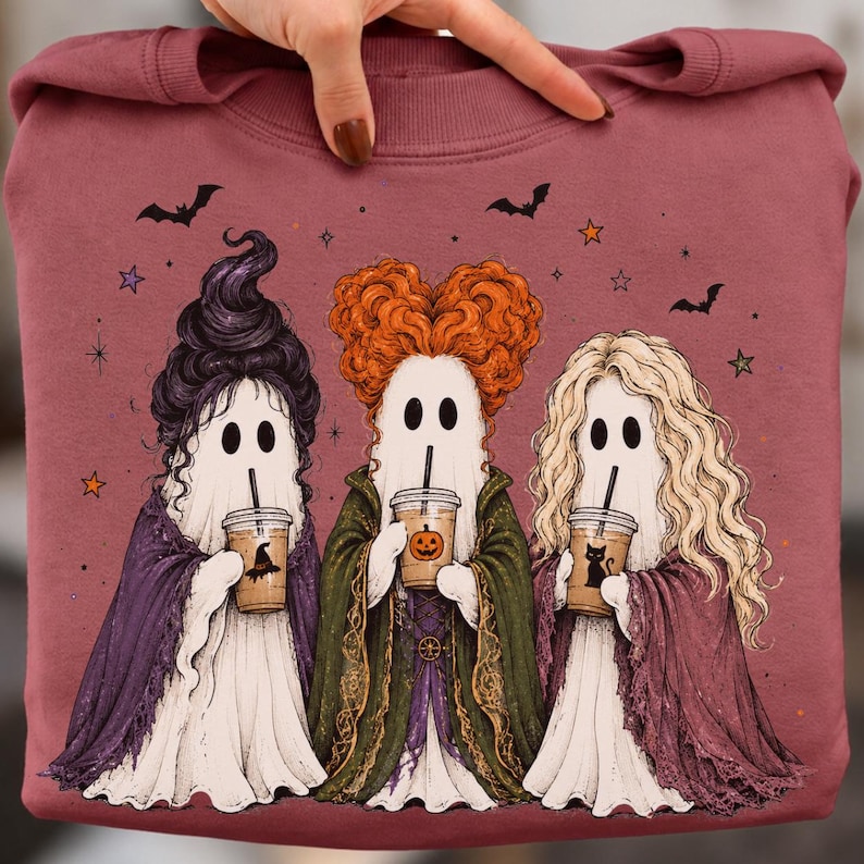 May include: A mauve sweatshirt featuring a graphic of three ghosts dressed as witches with distinct hairstyles, each holding an iced coffee cup. The ghosts are adorned with purple, green, and maroon capes, surrounded by small bats and stars.