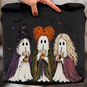 May include: A black sweatshirt featuring a graphic of three ghosts dressed as witches, each holding a plastic cup with a straw. The ghosts have distinct hairstyles in purple, orange, and blonde, and wear capes in purple, green, and maroon.