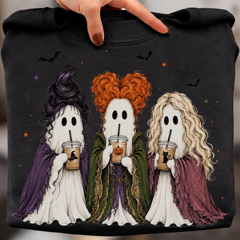 May include: A black sweatshirt featuring a graphic of three ghosts dressed as witches, each holding a plastic cup with a straw. The ghosts have distinct hairstyles in purple, orange, and blonde, and wear capes in purple, green, and maroon.