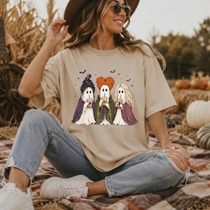 May include: A beige short-sleeve t-shirt featuring a graphic print of three ghosts dressed as witches, each holding a coffee cup. The ghosts have distinct hairstyles and colored cloaks in purple, green, and maroon, surrounded by small bats and stars.