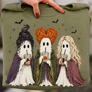 May include: An olive green sweatshirt featuring a graphic of three ghosts dressed as witches. Each ghost has distinct hair, wears a colored cloak, and holds an iced coffee cup. The design includes small bats, stars, and a pumpkin icon on one cup.