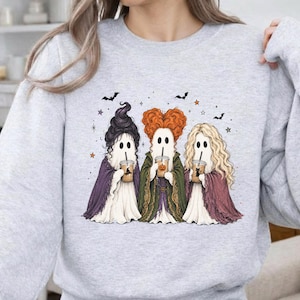 May include: A light gray crewneck sweatshirt featuring a graphic of three ghosts dressed as witches, each holding a coffee cup. The ghosts have distinct hairstyles in black, orange, and blonde, and are surrounded by small stars and bats.