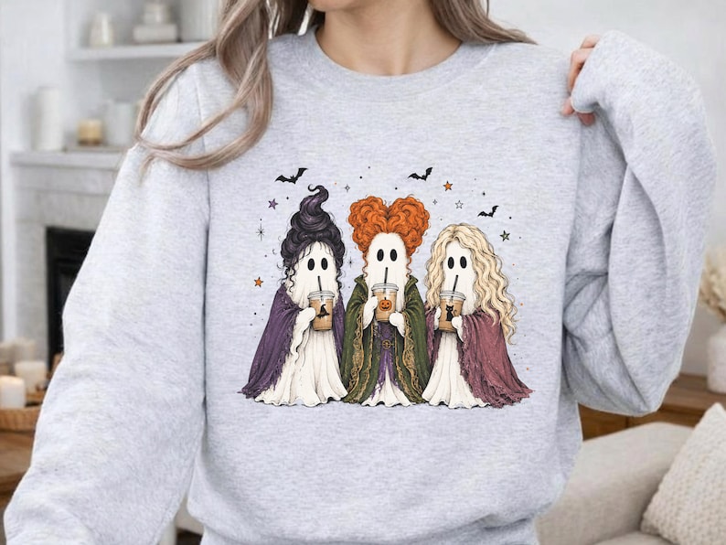 May include: A light gray crewneck sweatshirt featuring a graphic of three ghosts dressed as witches, each holding a coffee cup. The ghosts have distinct hairstyles in black, orange, and blonde, and are surrounded by small stars and bats.