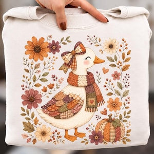 Könnte beinhalten: Ein cremefarbenes Sweatshirt mit einer Grafik einer Ente, die eine karierte Schleife und einen passenden Schal trägt. Die Ente ist von herbstlichen Blumen, Blättern und einem Kürbis umgeben. Das Design verwendet warme Orange- und Brauntöne.