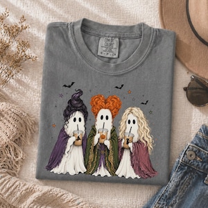 May include: A charcoal gray t-shirt featuring a graphic of three ghosts dressed as witches with distinct hairstyles, each holding a plastic cup with a straw. The ghosts wear purple, green, and maroon capes. Small bats and stars surround them.