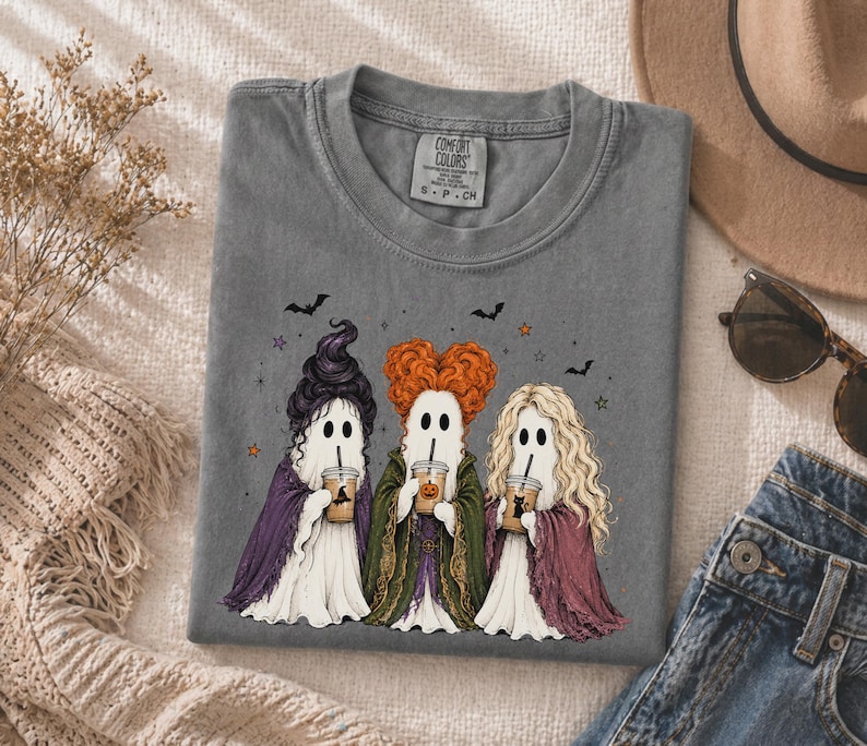 May include: A charcoal gray t-shirt featuring a graphic of three ghosts dressed as witches with distinct hairstyles, each holding a plastic cup with a straw. The ghosts wear purple, green, and maroon capes. Small bats and stars surround them.