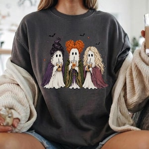 May include: A charcoal gray crewneck sweatshirt featuring a graphic of three ghosts dressed as witches, each holding a coffee cup. The ghosts have distinct hairstyles in purple, orange, and blonde, and are surrounded by small black bats and stars.