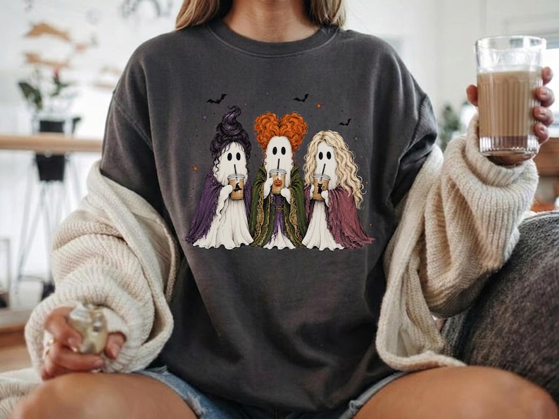 May include: A charcoal gray crewneck sweatshirt featuring a graphic of three ghosts dressed as witches, each holding a coffee cup. The ghosts have distinct hairstyles in purple, orange, and blonde, and are surrounded by small black bats and stars.