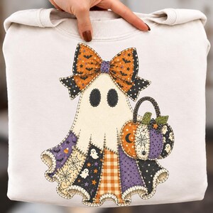 Könnte beinhalten: Ein cremefarbenes Sweatshirt mit einer Grafik eines Geistes, der eine große orange-schwarze Schleife trägt. Der Körper des Geistes ist im Patchwork-Stil gestaltet und zeigt verschiedene Halloween-Motive wie Spinnennetze, Sterne und kleine Geister.