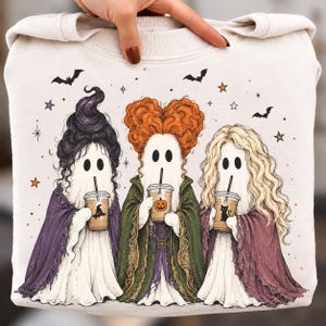 Puede incluir: Una sudadera de color crema con un gráfico de tres fantasmas vestidos de brujas. Cada fantasma tiene un peinado distinto, lleva una capa de color y sostiene un vaso de plástico con pajita. El diseño incluye murciélagos, estrellas e iconos.