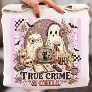 Può includere: Una felpa color crema con la grafica di due fantasmi che guardano una televisione vintage che mostra un gatto nero. Il design include un teschio, una cassetta, un taccuino con scritto Case Notes, popcorn e il testo True Crime & Chill.
