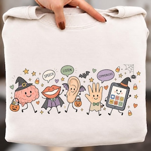Könnte beinhalten: Ein cremefarbenes Sweatshirt mit Rundhalsausschnitt und einer bunten Halloween-Grafik, die ein Gehirn, Lippen, ein Ohr, eine Hand und ein Tablet zeigt. Die Figuren tragen Hexenhüte und Umhänge und halten Süßigkeiten-Eimer. Sprechblasen enthalten die Wörter SPEECH, LISTEN und COMMUNICATE.